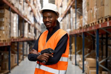 Güvenilir Afro-Amerikan ambar işçisi elinde barkod tarayıcısı ile lojistik merkezinde kolları bağlı gülümsüyor.