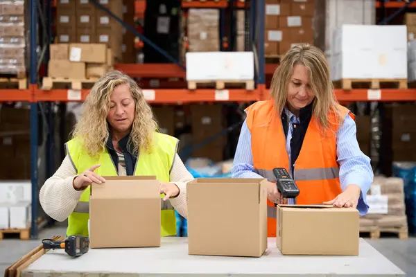 İki kadın depo çalışanı bir lojistik merkezinde karton kutular hazırlıyor, kodları kontrol ediyor ve tarıyor.