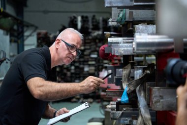 Deneyimli metal işçisi özel mekanik parçaları gösterirken panoyu tutuyor ve metalurji kumaşında kalite kontrolü yapıyor.