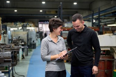 İki fabrika işçisi gülümsüyor ve bir metal fabrikasında üretim planlarını tartışıyor.