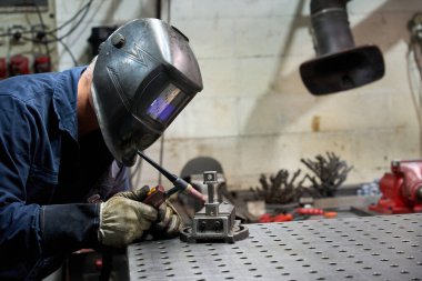 Metal işçisi koruyucu maske takıyor. Metalurji atölyesinde özel bir mekanik parça kaynak yapıyor.