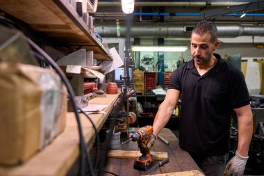 Metal işçisi yoğun bir atölyede özel mekanik parçalar yaparken kablosuz matkabı dikkatlice kullanarak endüstriyel üretimi gösteriyor.
