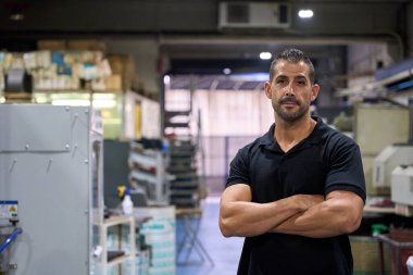 Bir fabrikada çapraz silahlarla duran kendinden emin bir metal işçisinin portresi endüstriyel çevrede uzmanlık ve gurur gösteriyor.