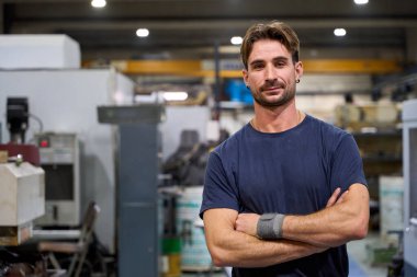 Bir fabrika atölyesinde çapraz silahlarla poz veren kendinden emin endüstriyel mühendis, metalurji üretiminde uzmanlık ve profesyonellik sergiliyor.