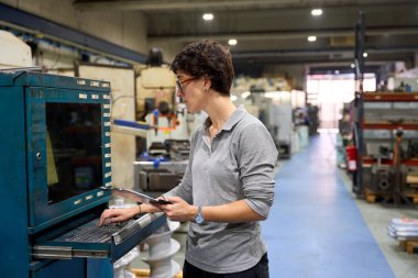 Bilgisayar ve dijital tablet kullanan kadın mühendis, özel mekanik parçalar üreten bir metalurji fabrikasında endüstriyel makineleri yönetiyor.