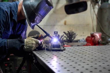 Koruyucu maske takan ve eldiven takan metal işçisi atölyedeki bir kaynakçıyı kullanarak özel mekanik bir parça kaynak yapıyor.