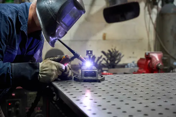 Koruyucu maske takan ve eldiven takan metal işçisi atölyedeki bir kaynakçıyı kullanarak özel mekanik bir parça kaynak yapıyor.