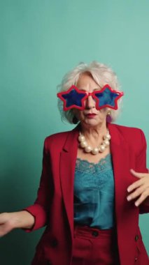 Kırmızı elbiseli iş kadını turkuaz arka planda farklı pozlar veriyor, yıldız şeklinde güneş gözlüğü takıyor.