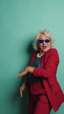 Kırmızı takım elbiseli, mavi gözlükle son sınıf güzeli dans ediyor ve turkuaz bir arka planda vuruşlar yapıyor.