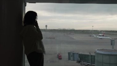 Gezgin, büyük bir pencereden havaalanı pistindeki faaliyetleri izlerken bir telefon görüşmesi yapıyor.