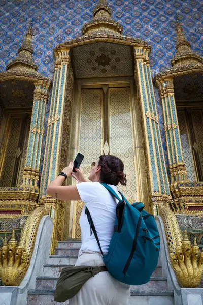 Sırt çantalı genç bir kadın gezisi sırasında Bangkok, Tayland 'daki büyük sarayın görkemli altın kapısının fotoğraflarını çekiyor.