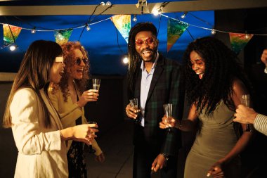 Çatı katındaki bir partide gülen ve şampanya içen çok ırklı arkadaşlar. Telli ışıklar ve renkli bayraklar altında.