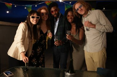 Çeşitli arkadaş grupları çatıda eğleniyor, ellerinde şampanya bardakları ve kutlama ışıklarının altında birlikte kutluyorlar.