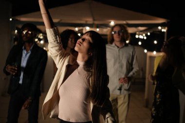 Genç bir kadın gece partisinden hoşlanıyor, kollarını kaldırarak dans ediyor, çok ırklı arkadaşlar arasında,