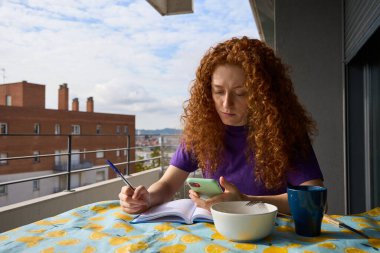 Serbest çalışan kadın balkonda çalışıyor not alıyor ve kahvaltıda akıllı telefonuna danışıyor.