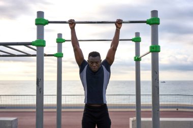 Odaklanmış genç siyah atlet şafak vakti deniz kenarındaki bir barfiks çubuğunda jimnastik yapıyor. Güç, adanmışlık ve sağlıklı bir yaşam tarzı sergiliyor.