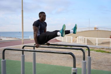 Kaslı siyah adam dışarıda antrenman yapıyor. Paralel barlarda oturuyor. Sahildeki bir arka planda güç ve zindelik gösterisi yapıyor.