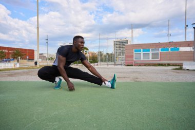 Konsantre sporcu, şehir parkındaki yeşil yapay çim üzerinde bacak esnetme hareketleri yapıyor, fitness ve kentsel çalışma yaşam tarzını destekliyor.