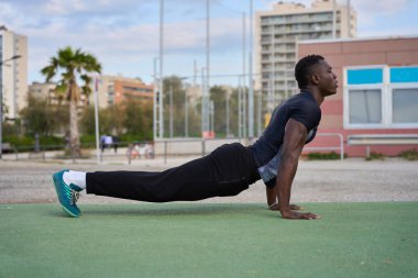 Atletik siyah adam açık havada antrenman yapıyor, yeşil bir yüzey üzerinde sahte tahta şınav çekiyor.