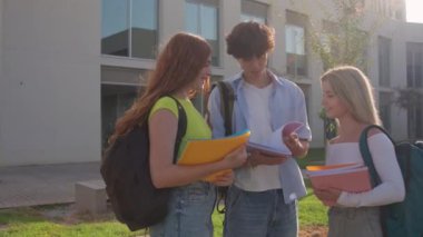 Üç üniversite öğrencisi güneşli bir kampüste birlikte notları tartışıyor ve gözden geçiriyorlar, işbirliği ve arkadaşlığın tadını çıkarıyorlar.