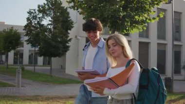 İki öğrenci kampüste yürüyor, ödevleri tartışıyor ve güneşli gökyüzünün altında güzel havanın tadını çıkarıyorlar.