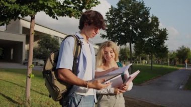 Yeşillik ve modern mimari ile çevrili bir kampüs yolunda dururken iki üniversite öğrencisi ders çalışıyor ve ödevlerini tartışıyorlar.