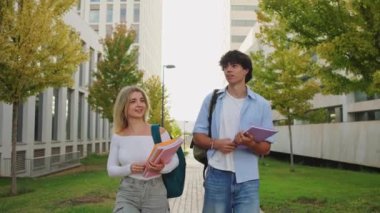 İki üniversite öğrencisi kampüs yolunda yürümekten keyif alıyor, kitap taşıyor ve sohbet ediyorlar.