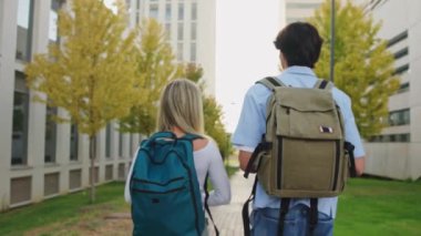Sırt çantalı iki üniversite öğrencisi kampüste yürüyüp konuşuyorlar, birbirleriyle eğleniyorlar.