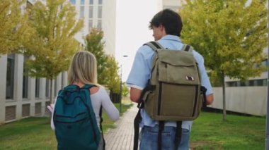 Sırt çantalı iki üniversite öğrencisi kampüste yürüyor ve dersler arasında sohbet ediyorlar.