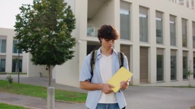 Üniversite öğrencisi elinde sarı bir dosya, güneşli bir günde kampüs binasının önünde poz verirken gülümsüyor.