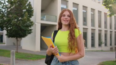 Üniversite öğrencisi kampüste klasörleri tutarken gülümsüyor, açık hava ortamında mutluluk ve güven yayıyor.