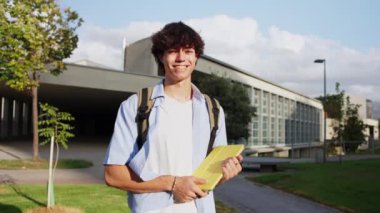Sırıtkan üniversite öğrencisi kampüs binasının dışında yürüyor, sarı defter taşıyor, güneşli bir günün tadını çıkarıyor, öğrenme sevincini ve olumluluğunu dile getiriyor.