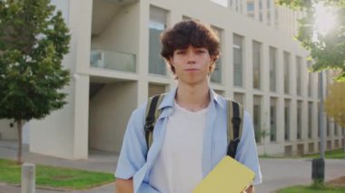 Sırt çantası ve kitapları olan genç bir adam güneşli bir üniversite kampüsünde neşeli akademik atmosferin tadını çıkararak gülümsüyor.