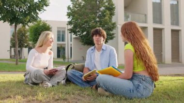 Üç üniversite öğrencisi neşeli bir sohbete dalarken güneşli kampüs çimlerinin keyfini çıkarıyorlar, kahkahalar ve arkadaşlıkları paylaşıyorlar.