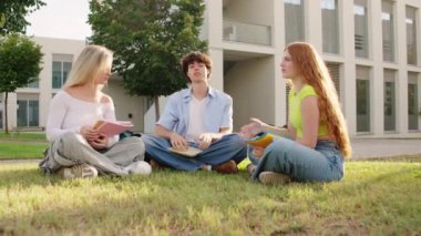 Üç üniversite öğrencisi, derslerine dalmış, yemyeşil kampüs çimlerinde dinamik bir sohbete girişiyor.