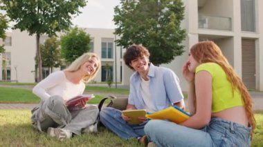 Çeşitli üniversite öğrencileri çimlerin üzerinde oturuyor, güneşli bir günde birlikte gülüyor ve ders çalışıyorlar, dostluk ve akademik işbirliği sergiliyorlar.