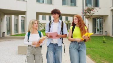 Üç üniversite öğrencisi kampüste güneşli bir günün tadını çıkarırken dışarıda sohbet edip kitap taşıyorlar.