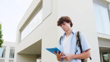 Üniversite öğrencisi modern kampüs binasının dışında yürürken bir defter okuyor. Akademik ortamda güneşli bir gün