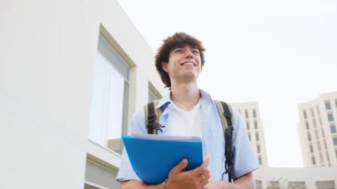 Sırt çantası ve klasörleri olan genç bir adam üniversite kampüsünde güvenle yürüyor, öğrenci hayatının ruhunu somutlaştırıyor.