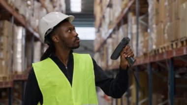 Ağır çekim sahnesinde güvenlik donanımı takmış bir depo çalışanının envanter taraması ve yoğun bir lojistik merkez koridorunda hassasiyet sağlaması.