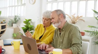 Olgun bir çift mutfak masasında oturmuş, faturalarla boğuşurken finansal durumlarını kontrol etmek için dizüstü bilgisayar kullanıyor.