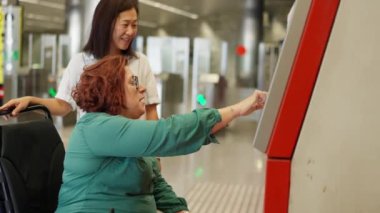 Tekerlekli sandalyedeki kadın bilet otomatını kullanırken, arkadaşı da toplu taşıma araçlarında yardım ve destek sağlıyor.
