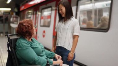 Genç bir kadın, metroda beklerken arkadaşını tekerlekli sandalyede teselli ediyor. Kahkaha ve desteği paylaşıyor.
