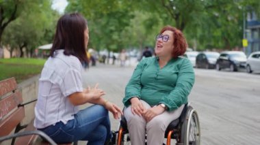 Kadın bankta oturuyor ve tekerlekli sandalye kullanan arkadaşının günü hakkında konuşmasını dinliyor.