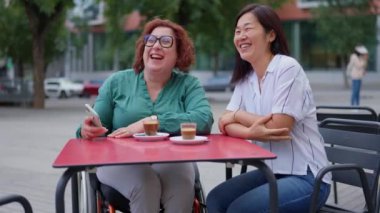 İki kadın, biri tekerlekli sandalye kullanıyor, selfie çekiyor ve açık bir kafede birlikte gülüyorlar.