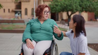 Tekerlekli sandalyedeki kadın, açık havada bir arkadaşıyla canlı bir sohbete girişiyor, dostluk ve bağlarını gösteriyor.