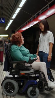 İki kadın gülüşüp sohbet ederken, biri diğerini metro istasyonunda tekerlekli sandalyeye itiyor.