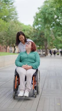 Bir kadın tekerlekli sandalyede gülen bir arkadaşını güneşli bir park yolunda itiyor. Doğada dostluk ve destek gösterisi yapıyor.