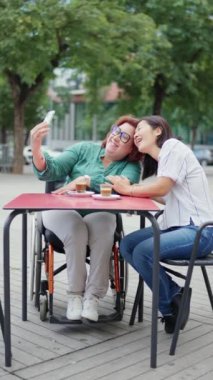 İki bayan arkadaş, biri tekerlekli sandalye kullanıyor, açık bir kafede kahve içerken selfie çekiyorlar.