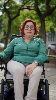 Tekerlekli sandalyede oturan gülümseyen kadın yeşil ağaçlar ve doğa ile çevrili bir parkta güneşli bir günün tadını çıkarıyor.